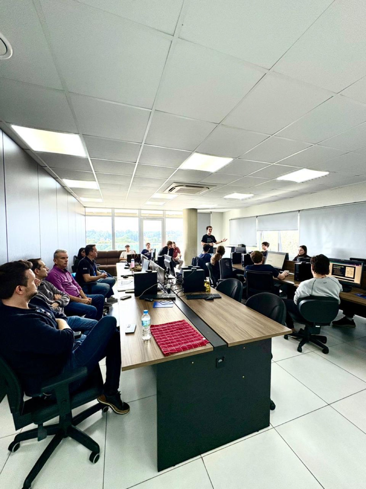 Residente apresentando para a equipe do CRS reunida no laboratório, com professores e colegas assistindo.