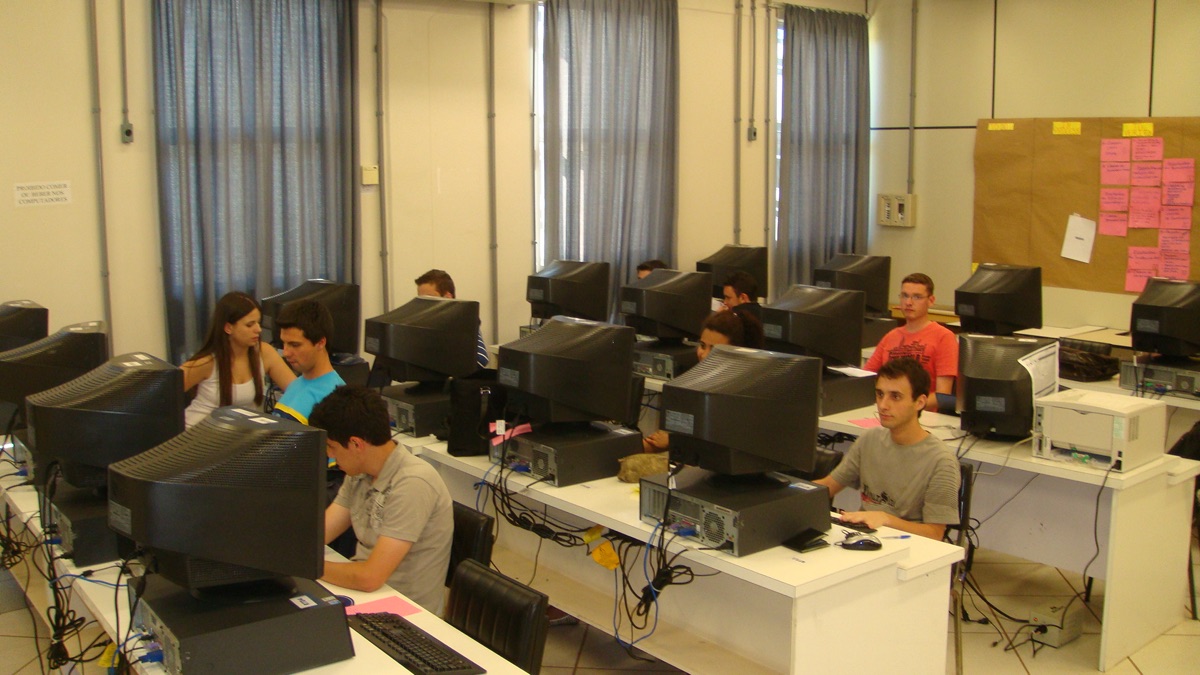 Residentes trabalhando em laboratório com monitores de tubo e um quadro de tarefas ao fundo.